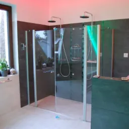 Nowoczesna łazienka z dwoma przeszklonymi kabinami prysznicowymi typu walk-in, wykończona ciemnoszarą płytką i podświetleniem LED, widok zza niskiej ścianki działowej.