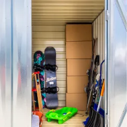 Wnętrze blaszanej komórki z deskami snowboardowymi Burton, nartami, sankami i kartonami, podłoga wyłożona drewnem.