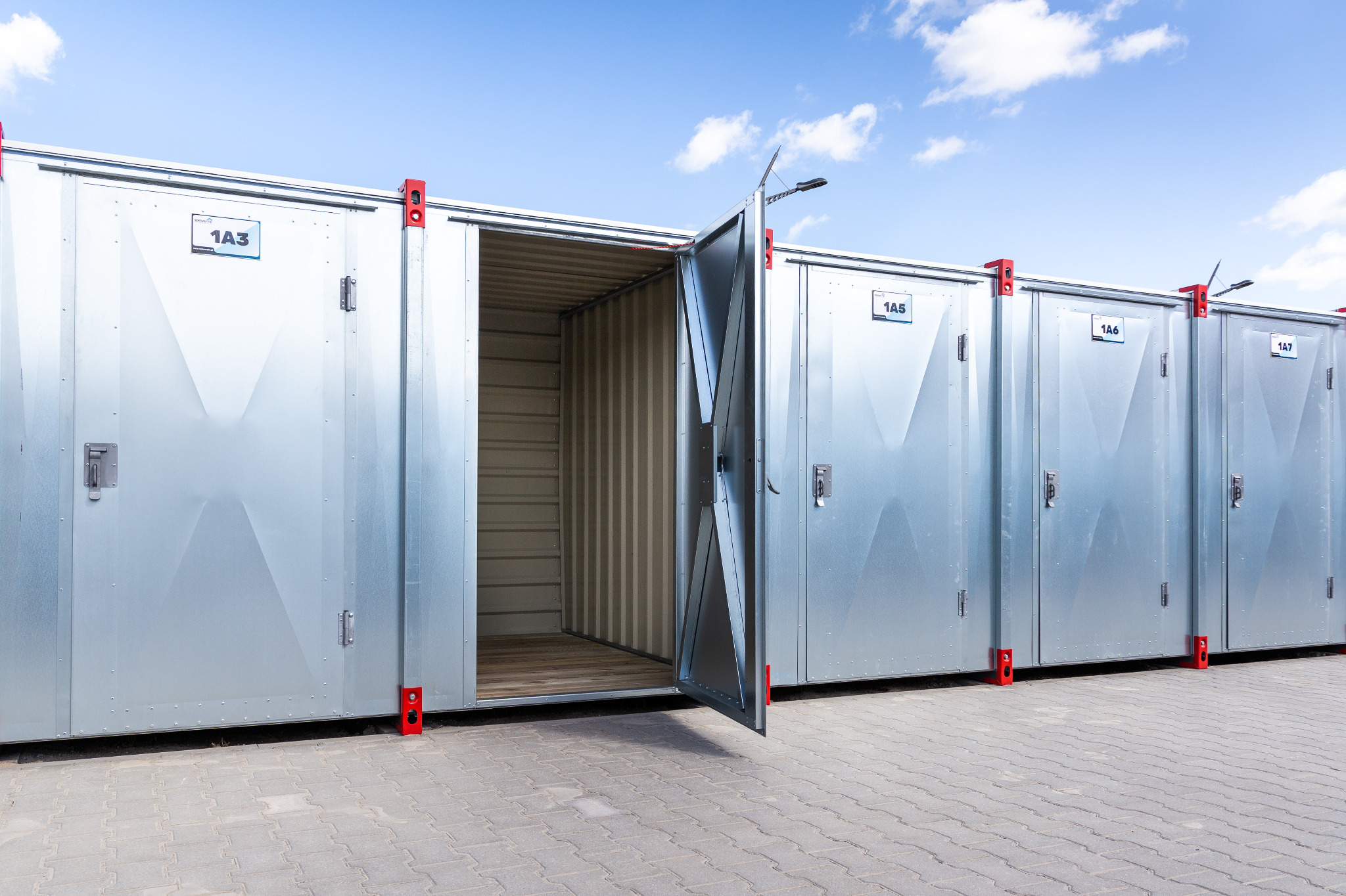 Rząd srebrnych kontenerów magazynowych z czerwonymi elementami mocującymi, jeden kontener z otwartymi drzwiami, widoczne wnętrze z pofalowanej blachy.