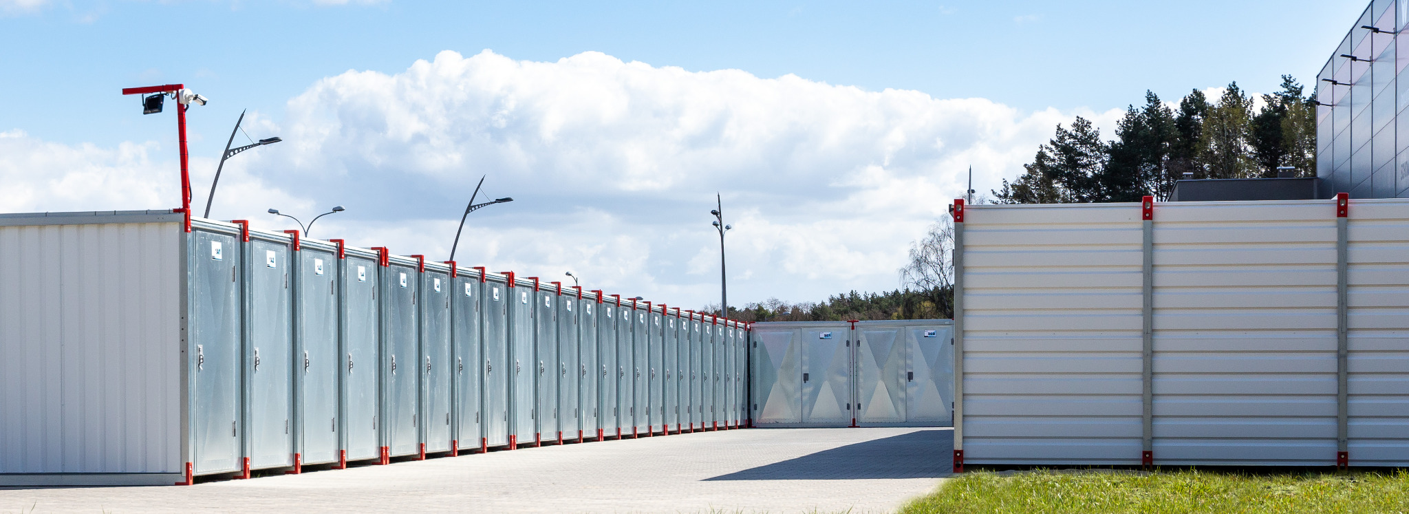 Rząd metalowych schowków samoobsługowych z czerwonymi akcentami, zewnętrzny monitoring w tle, betonowy plac i fragment trawnika.