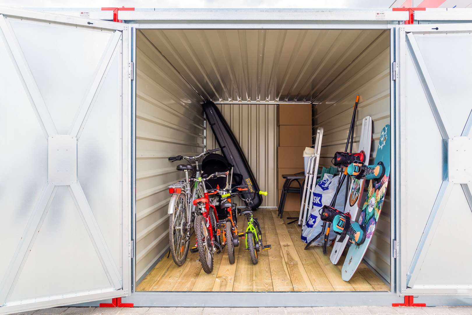 Wnętrze otwartego kontenera magazynowego z trzema rowerami (damski, dziecięcy, mały rowerek), deskami do kitesurfingu, pudłami kartonowymi i akcesoriami sportowymi na drewnianej podłodze.