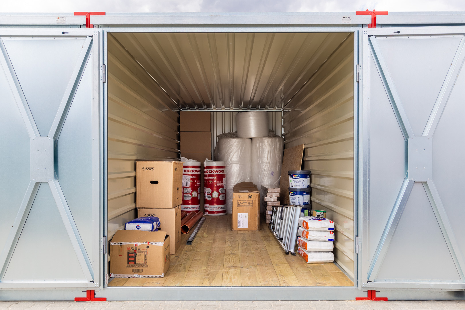 Otwarty kontener magazynowy z drewnianą podłogą, wypełniony kartonami, rolkami folii, wełną mineralną Rockwool, farbami Dulux i workami z zaprawą budowlaną.