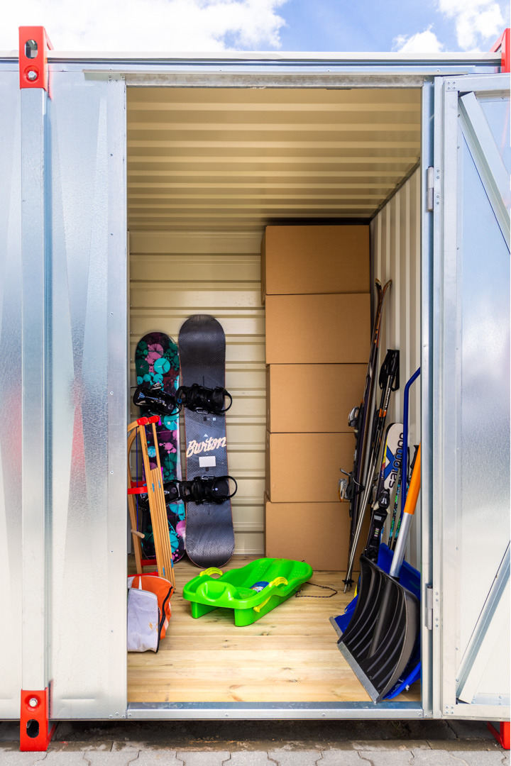 Wnętrze blaszanej komórki z deskami snowboardowymi Burton, nartami, sankami i kartonami, podłoga wyłożona drewnem.