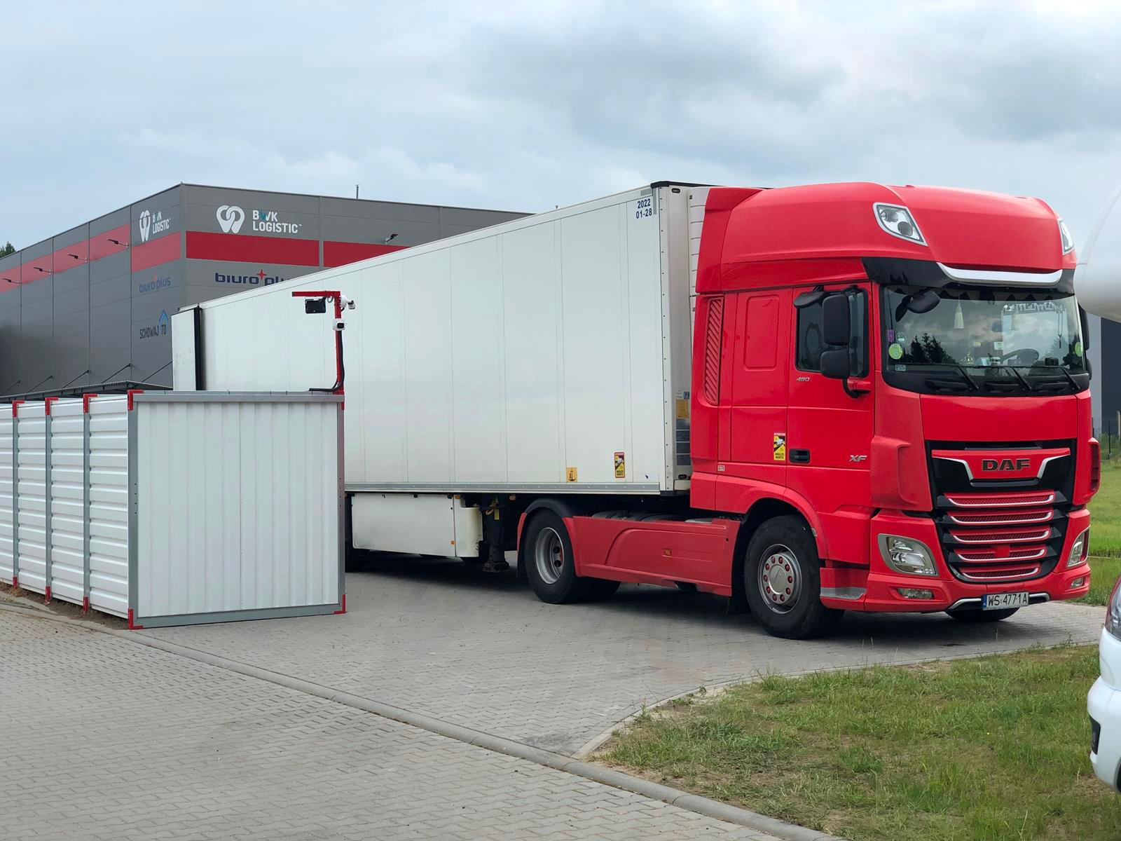 Czerwona ciężarówka DAF z białą naczepą stojąca obok białych, metalowych kontenerów magazynowych na tle budynku firmy logistycznej.