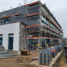 Budynek w trakcie prac, rusztowania pokryte czarnym materiałem izolacyjnym, widoczny pracownik w odblaskowej kamizelce, przygotowane materiały budowlane na ziemi, pochmurne niebo.