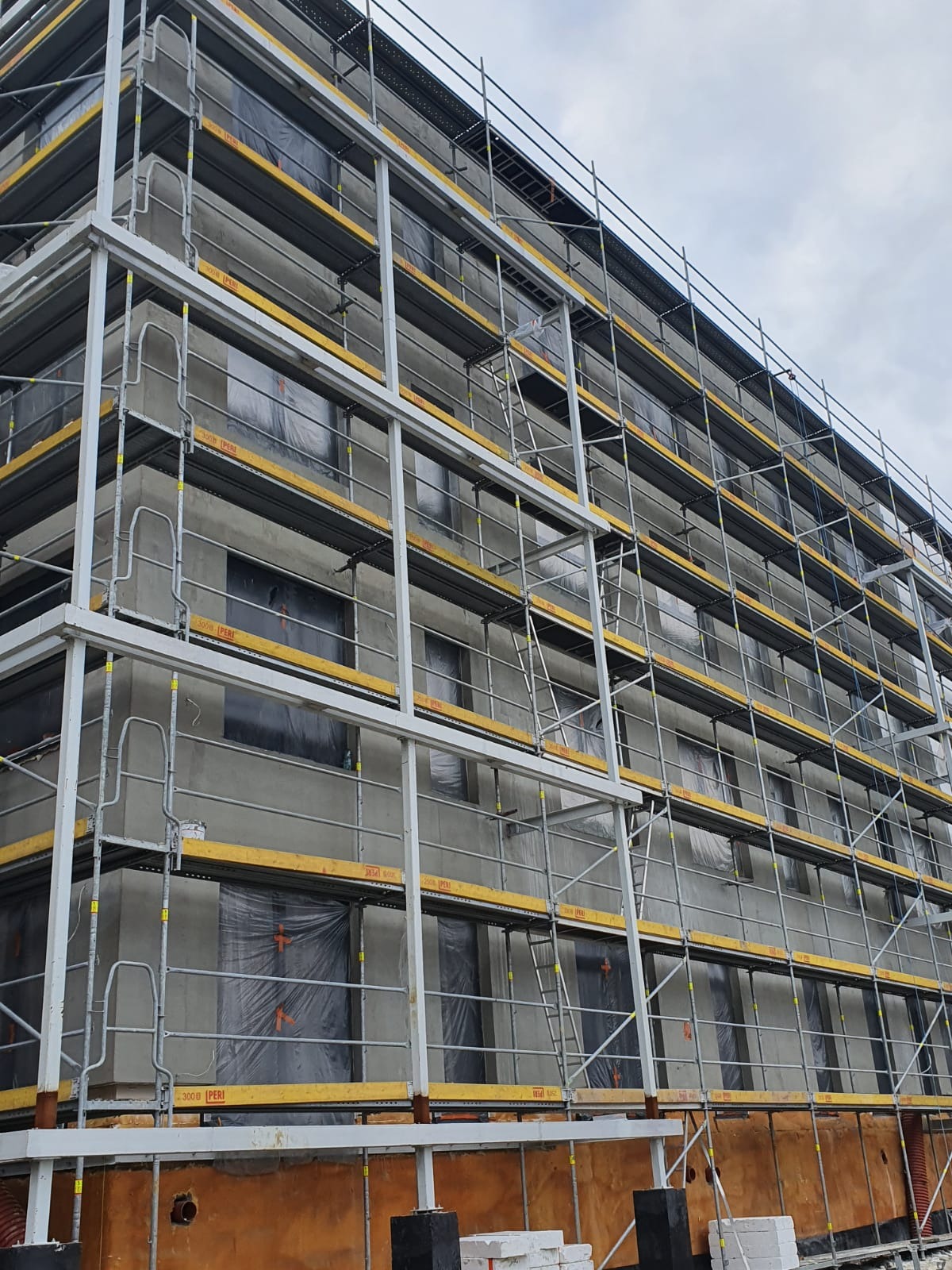 Budynek wielopiętrowy w trakcie prac elewacyjnych, otoczony rusztowaniem, okna zabezpieczone folią, widoczne materiały budowlane u podstawy.