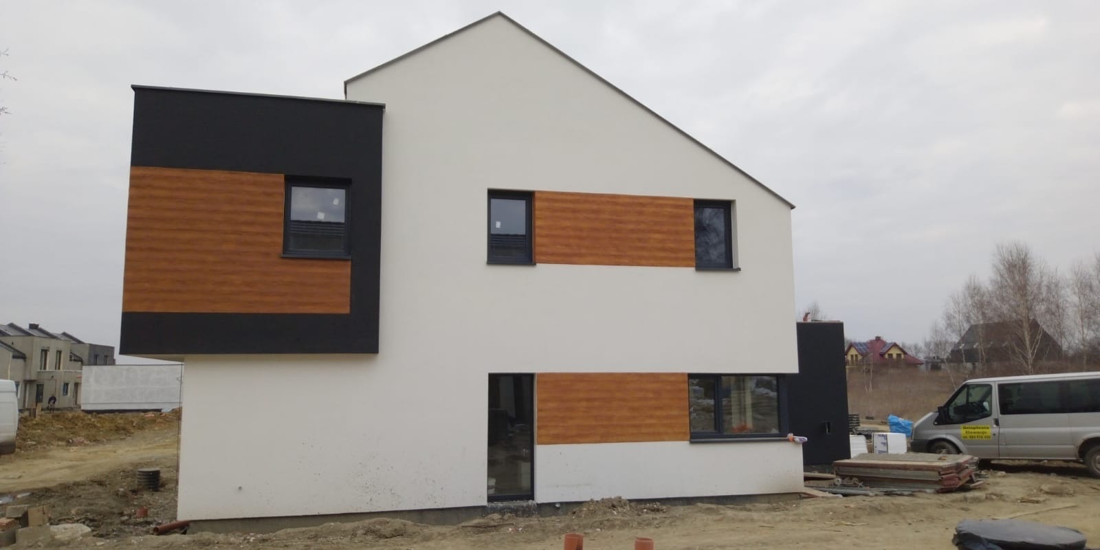 Nowoczesny dom jednorodzinny z białą elewacją, elementami dekoracyjnymi z paneli imitujących drewno i czarnymi ramami okiennymi, w trakcie prac wykończeniowych na zewnątrz.