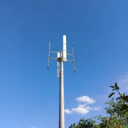 Pionowa turbina wiatrowa z trzema białymi skrzydłami i logo producenta na tle błękitnego nieba z lekkimi chmurami, widok od dołu z fragmentem zieleni w prawym dolnym rogu.