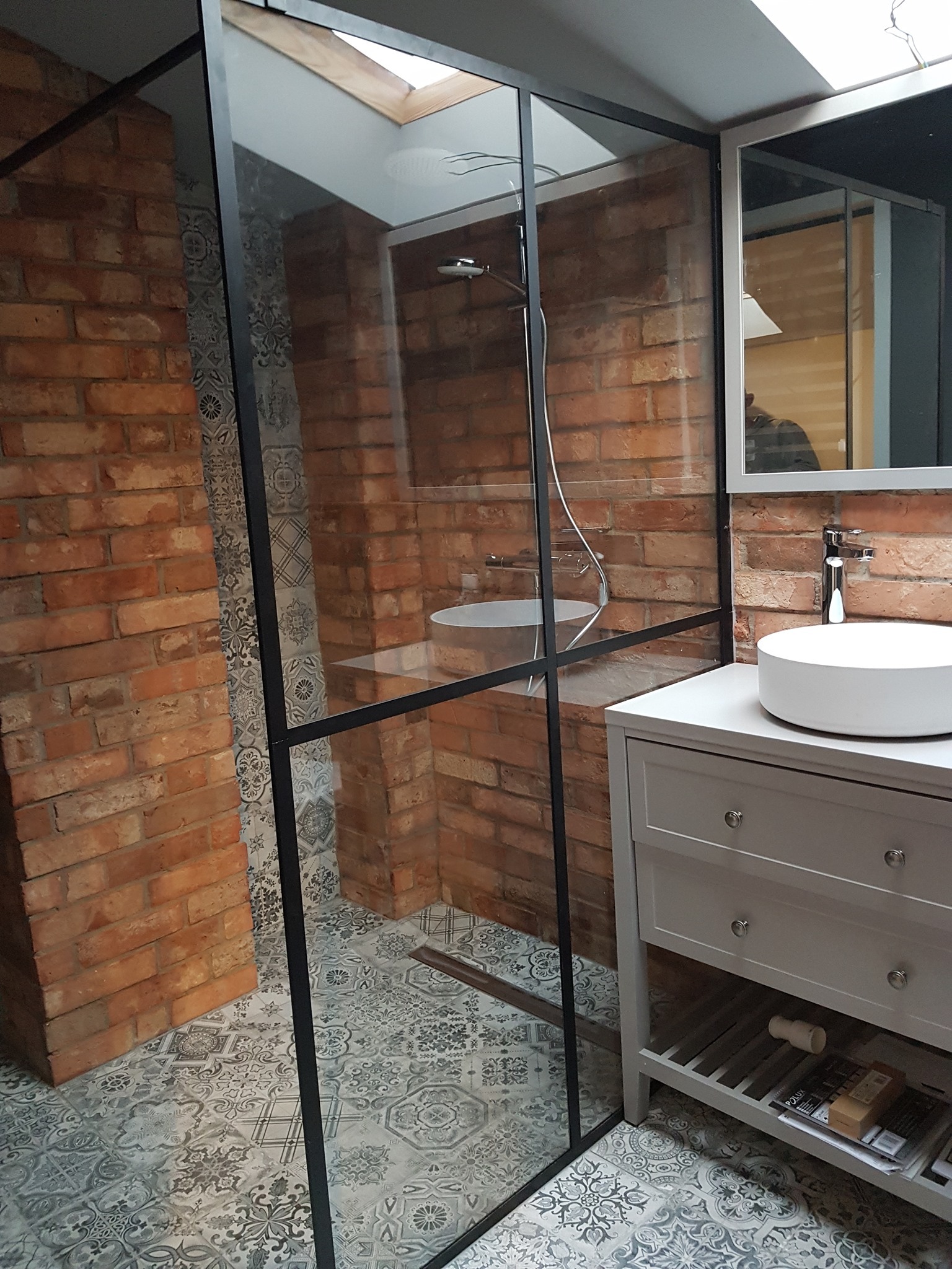 Nowoczesna łazienka z kabiną prysznicową typu walk-in, umywalką nablatową i ścianami z cegły, oświetlona światłem z okna dachowego.