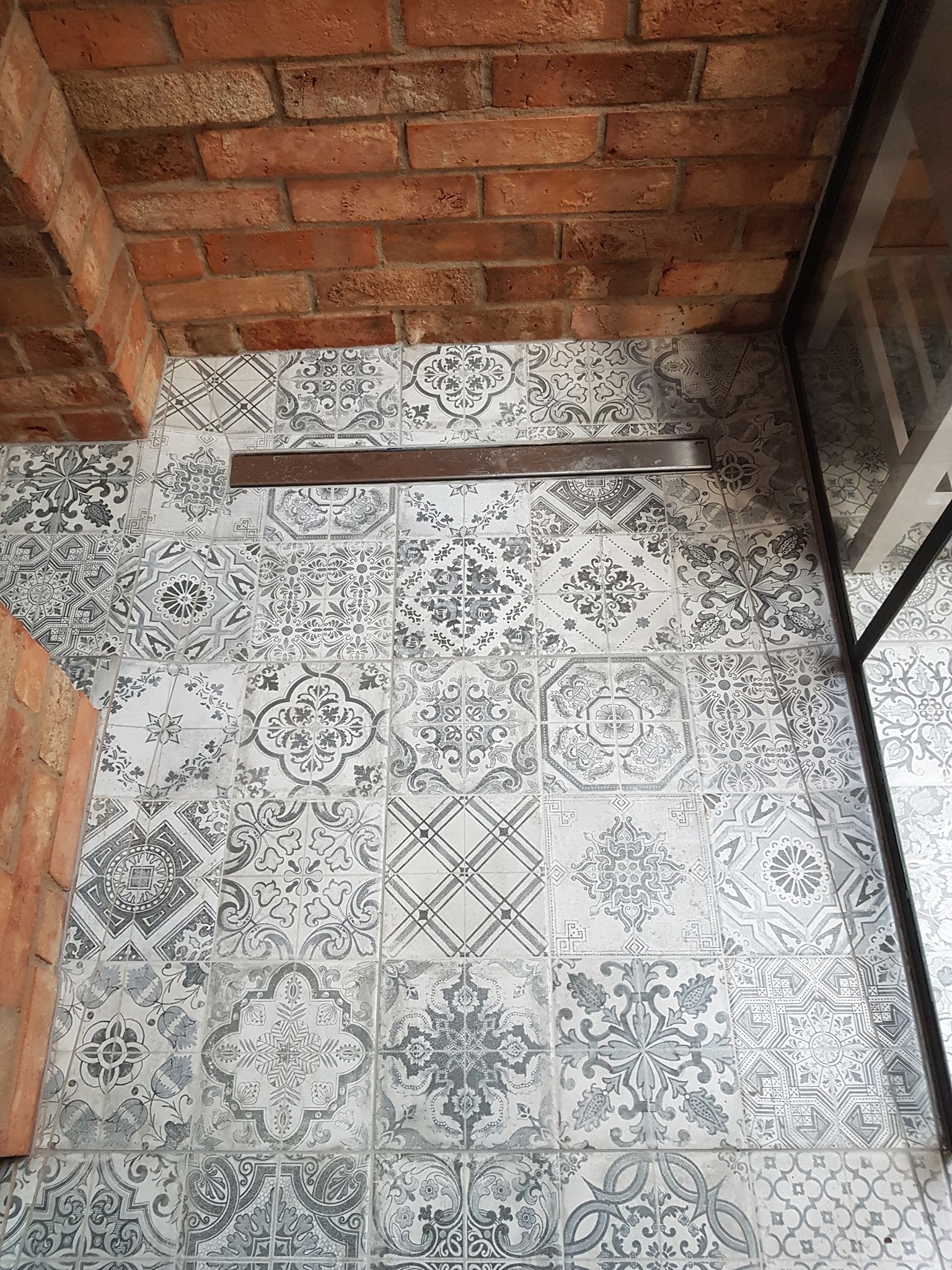 Wykończona podłoga łazienki z wzorzystych, szaro-białych płytek ceramicznych, z odpływem liniowym i fragmentem ściany z cegły oraz kabiny prysznicowej.