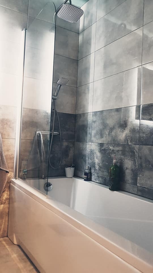 Wykończona łazienka z wanną obudowaną białym panelem, szklanym parawanem nawannowym i szaro-białymi płytkami imitującymi beton, z widoczną słuchawką prysznicową i deszczownicą.