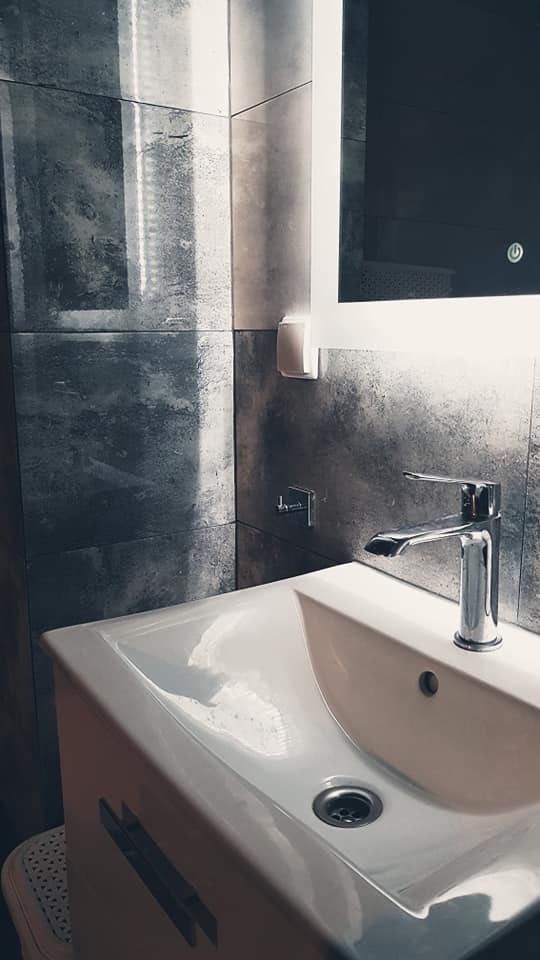 Nowoczesna łazienka z umywalką nablatową i baterią chromowaną, podświetlane lustro LED, szare płytki imitujące beton na ścianie, widoczny fragment taboretu pod umywalką.