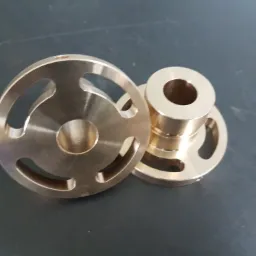 Dwa precyzyjnie obrobione, mosiężne elementy. Jeden w kształcie dysku z trzema wycięciami, drugi cylindryczny z kołnierzem z wycięciami, leżące na ciemnej, lekko zarysowanej powierzchni.