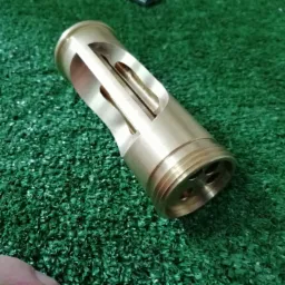 Mosiężny cylinder z precyzyjnymi wycięciami i otworami, leżący na zielonej sztucznej trawie, ukazujący detale obróbki CNC.