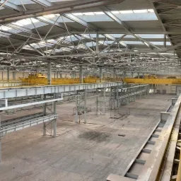 Wnętrze hali produkcyjnej w trakcie budowy, widoczne stalowe konstrukcje nośne, suwnice oraz oświetlenie dachowe.
