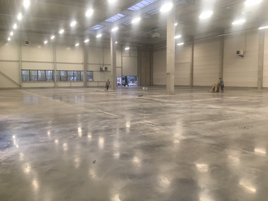 Duży, pusty magazyn z polerowaną betonową posadzką. Dwóch pracowników w oddali, jeden zamiata podłogę, drugi używa maszyny do polerowania. Światło dzienne wpada przez okna i świetliki dachowe.