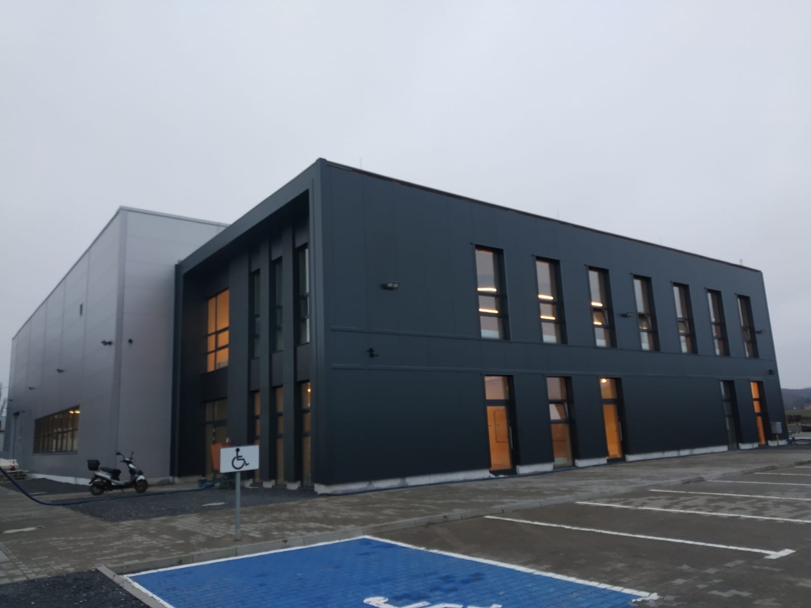 Nowoczesny budynek biurowy z szarą elewacją, widoczne miejsca parkingowe i skuter elektryczny zaparkowany obok znaku dla niepełnosprawnych.
