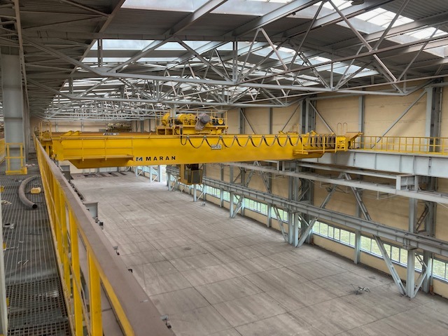 Żółty suwnicowy dźwig z napisem MARAN w hali produkcyjnej z widoczną konstrukcją stalową dachu i oknami w tle.