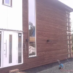 Elewacja domu z drewnianą okładziną w odcieniach brązu, białe okna i drzwi. Widoczne narzędzia i materiały budowlane sugerują etap wykończeniowy.