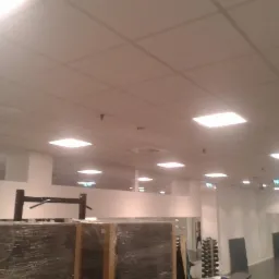 Montaż sufitu podwieszanego z oświetleniem w Alwerni. Widoczne płyty sufitowe i oprawy. Prace wykończeniowe w pomieszczeniu z lustrami i sprzętem sportowym.