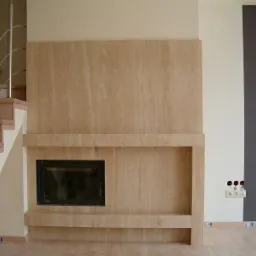 Nowoczesny kominek z jasnego trawertynu w salonie, z widocznymi schodami i elementami dekoracyjnymi ścian. Minimalistyczny design z akcentami drewna i metalu.