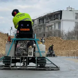 Pracownik w żółtej kamizelce obsługuje maszynę do zacierania betonu na budowie, w tle widoczny budynek w rusztowaniach i wykop ziemny zabezpieczony barierkami.