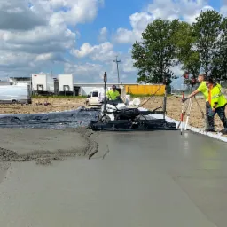 Dwóch pracowników w kamizelkach odblaskowych, używających niwelatora laserowego i maszyny do wygładzania betonu podczas wylewania nowej nawierzchni na placu budowy, z widocznymi budynkami w tle.