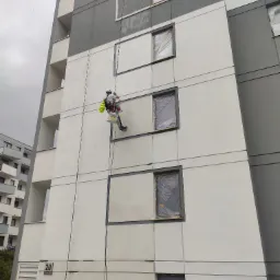 Pracownik na linach zabezpieczających, w trakcie odświeżania elewacji budynku wielorodzinnego, okna zabezpieczone folią, widoczne dwa wiadra z farbą.