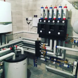 Kotłownia z kotłem Vaillant, zasobnikiem ciepłej wody i rozdzielaczem z manometrami, rury w kolorze szarym, instalacja po montażu, płytki na ścianach.