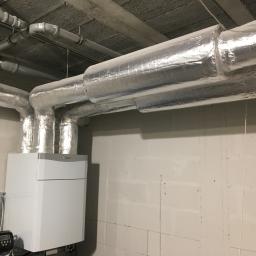 Hydro-Instal - Nowoczesny, biały piec gazowy marki Vaillant z podłączonymi, zaizolowanymi srebrną folią przewodami wentylacyjnymi, umieszczony w surowym pomieszczeniu z betonowym stropem i ścianami z bloków.