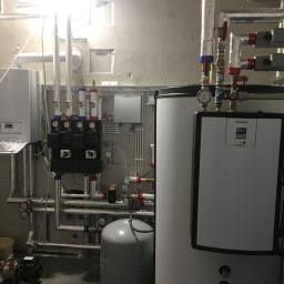 Hydro-Instal - Kotłownia z białym kotłem gazowym Vaillant, zbiornikiem ciepłej wody, pompą obiegową i licznymi rurami z zaworami, manometrami i izolacją termiczną, widoczne elementy instalacji grzewczej...