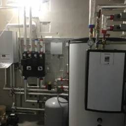 Kotłownia z białym kotłem gazowym Vaillant, zbiornikiem ciepłej wody, pompą obiegową i licznymi rurami z zaworami, manometrami i izolacją termiczną, widoczne elementy instalacji grzewczej...