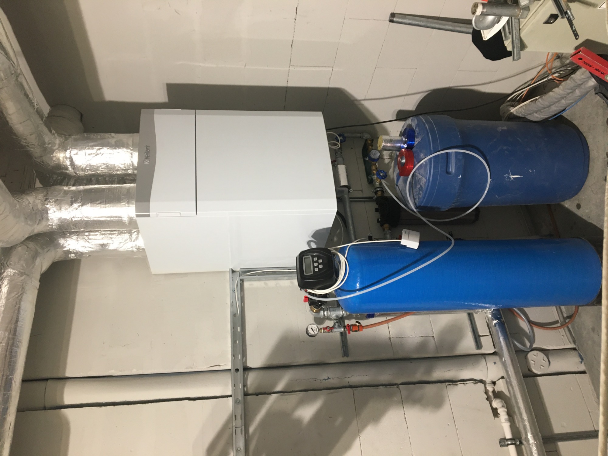 Kocioł gazowy Vaillant z podłączonymi rurami w izolacji aluminiowej, obok niebieskie zbiorniki na wodę i filtry, zamontowane na ścianie z bloków betonowych.