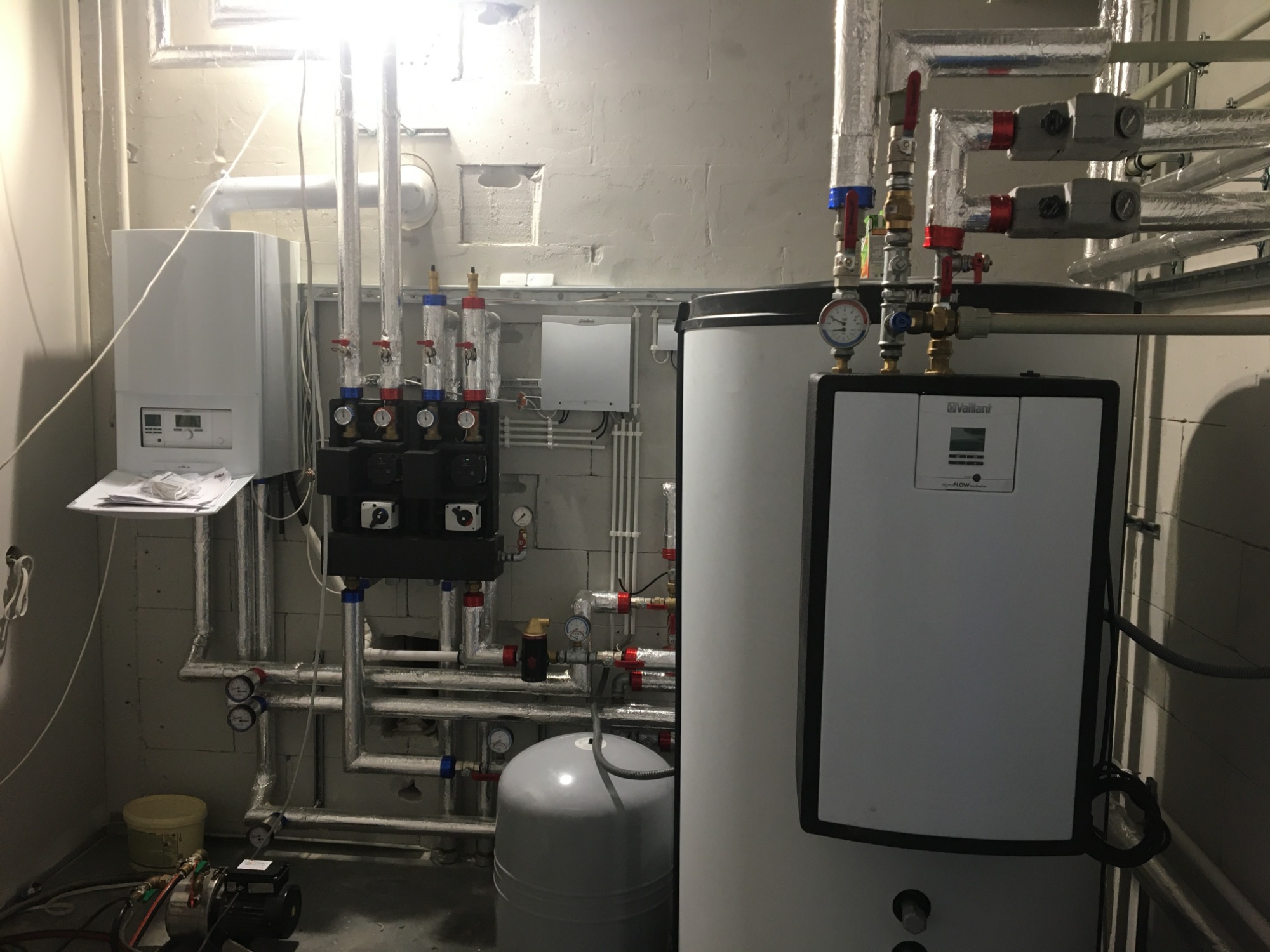 Kotłownia z białym kotłem gazowym Vaillant, zbiornikiem ciepłej wody, pompą obiegową i licznymi rurami z zaworami, manometrami i izolacją termiczną, widoczne elementy instalacji grzewczej...