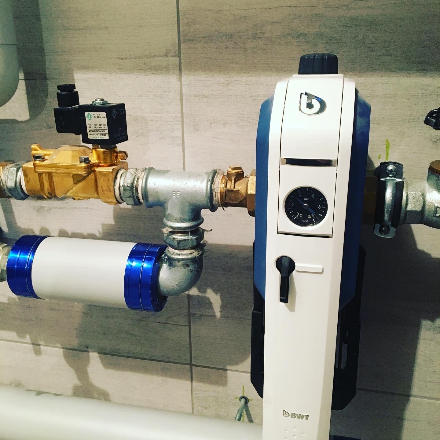 Filtr do wody BWT z manometrem i zaworem elektromagnetycznym, zainstalowany na rurach wodociągowych z szarymi płytkami w tle.
