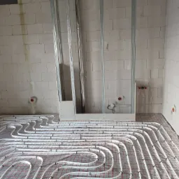 Montaż ogrzewania podłogowego w surowym wnętrzu w Sopocie, widoczne rury na folii z siatką, ściany z bloczków i metalowe profile.