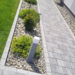 Ścieżka z szarej kostki brukowej prowadząca wzdłuż domu, obok rabaty z krzewami, kamykami i nowoczesnym oświetleniem.
