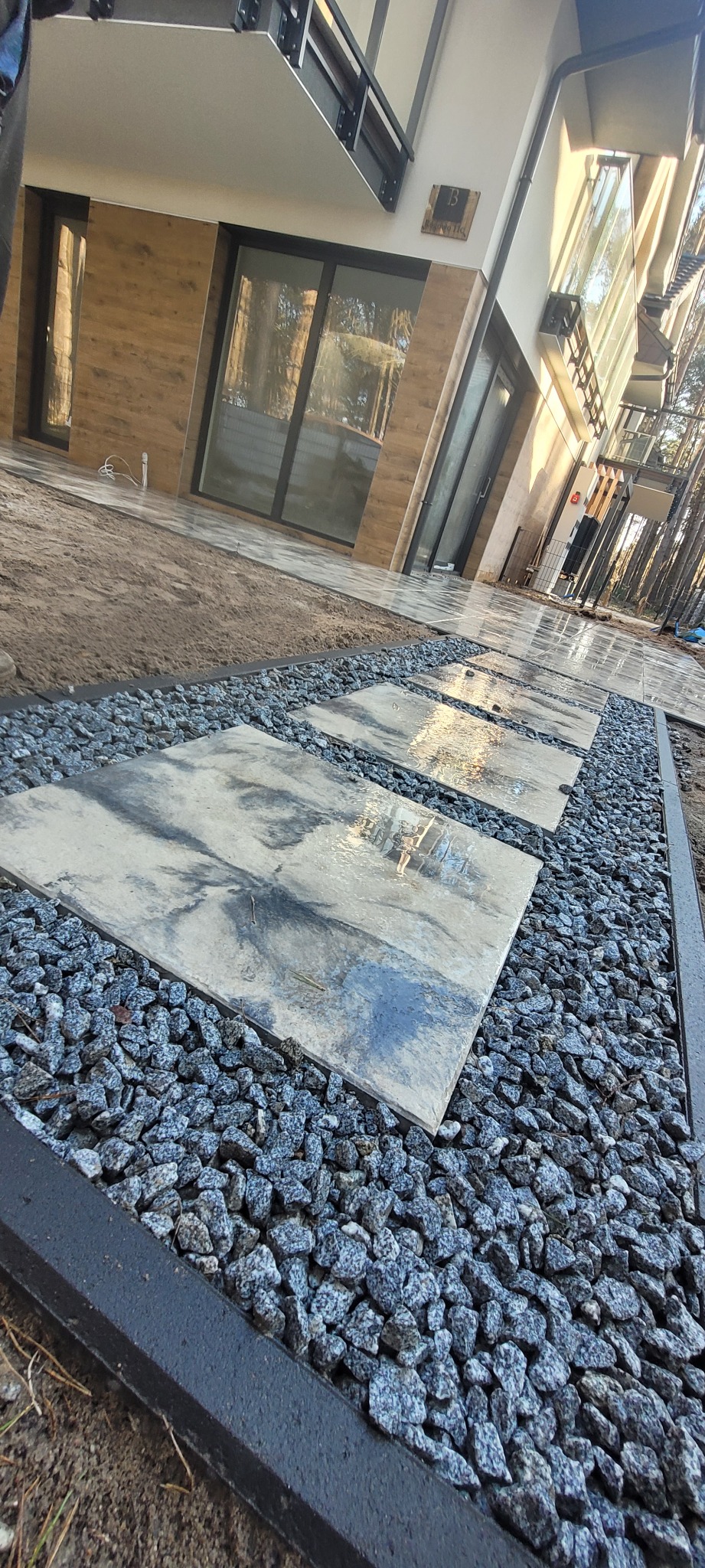 Ścieżka z płyt betonowych ułożona na szarym grysie granitowym, obrzeża z czarnego betonu, nowoczesny dom jednorodzinny w tle.