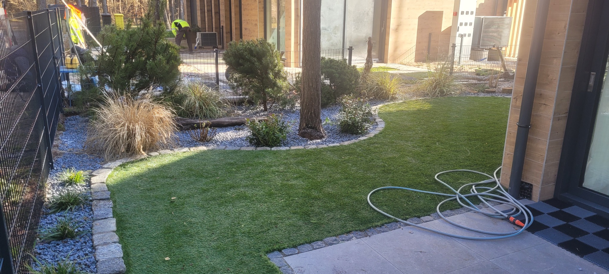 Ułożona kostka granitowa oddzielająca trawnik od rabat z ozdobnymi krzewami i trawami, widoczny wąż ogrodowy i pracownik w oddali.