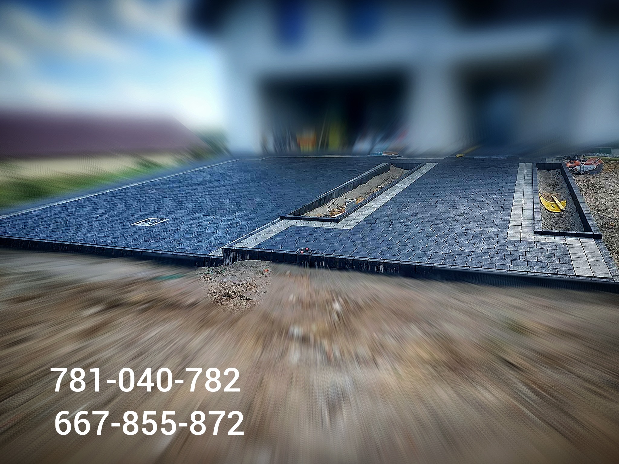 Fragment podjazdu z ułożoną ciemnoszarą kostką brukową, oddzielony pasami jasnej kostki, widoczne korytka odwadniające z piaskiem, w tle rozmazany budynek.