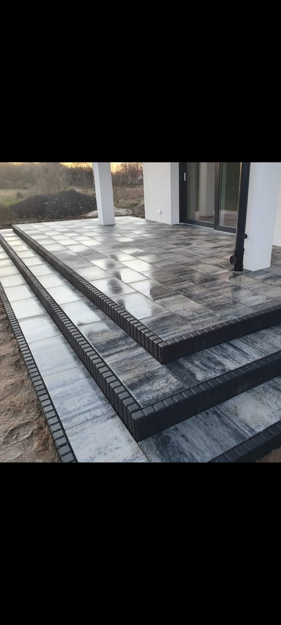Trzy stopnie schodów zewnętrznych wyłożone szarą kostką brukową z ciemnoszarym obrzeżem, prowadzące na taras z kostki, widok na białe filary i drzwi tarasowe, w tle hałda ziemi.