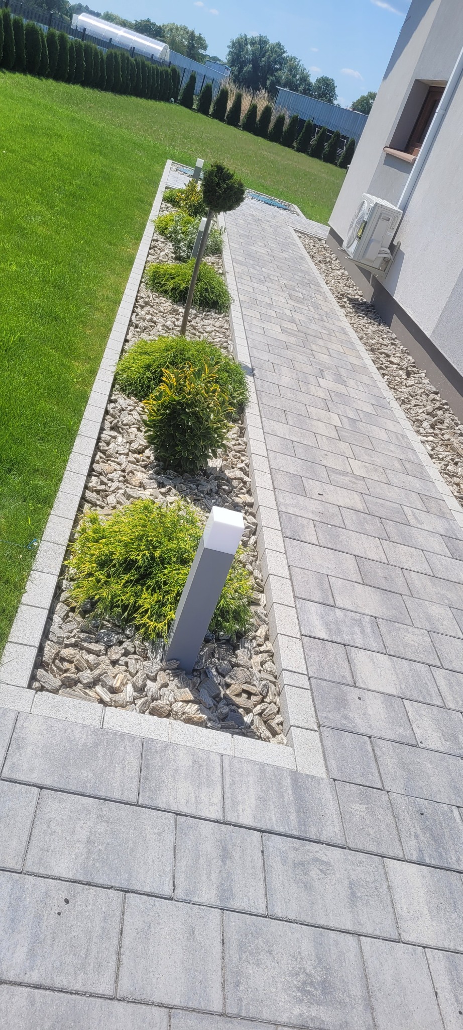 Ścieżka z szarej kostki brukowej prowadząca wzdłuż domu, obok rabaty z krzewami, kamykami i nowoczesnym oświetleniem.