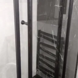 Szklana kabina prysznicowa z czarnymi profilami na tle białych i czarnych płytek imitujących marmur, widoczny grzejnik drabinkowy.