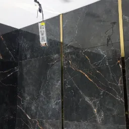 Czarne, marmurowe płytki ścienne z złotymi listwami wykończeniowymi w narożniku łazienki, widoczny fragment instalacji elektrycznej pod sufitem.