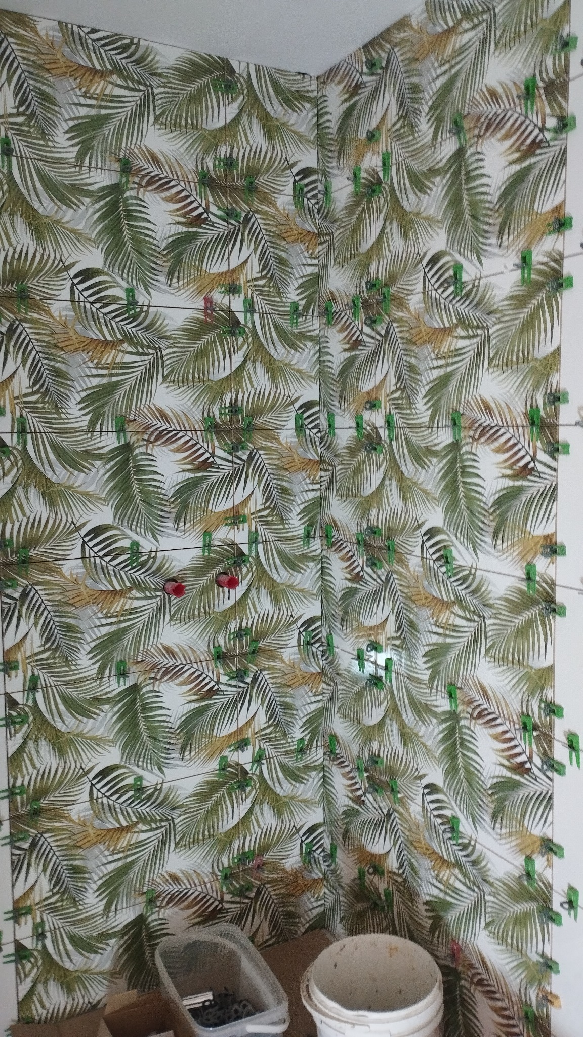 Ściana w trakcie układania płytek z motywem liści palmowych, z użyciem klipsów poziomujących, widoczny wiadro i pojemnik z akcesoriami do układania płytek.