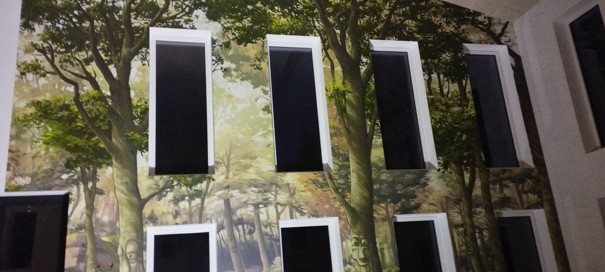 Ściana z fototapetą leśną i wkomponowanymi oknami z białymi ramami. Nietypowe połączenie natury i architektury wewnątrz pomieszczenia.