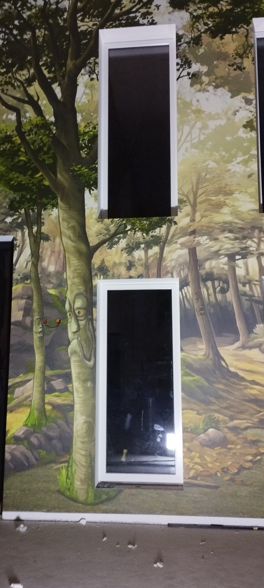 Dwa pionowe, prostokątne lustra z białą ramą na ścianie z fototapetą przedstawiającą las. Na tapecie widoczne drzewa z wyrazistymi twarzami.