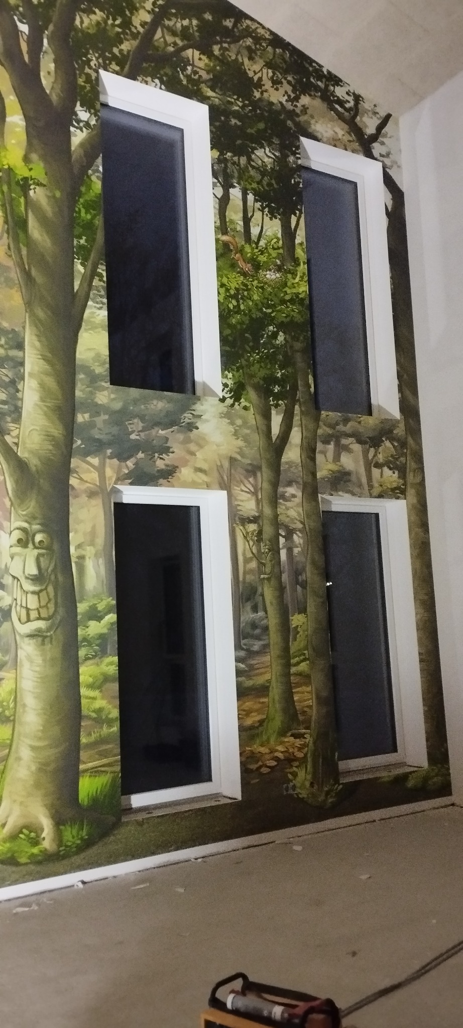 Ściana z fototapetą leśną i czterema oknami z białymi ramami. Na pierwszym planie narzędzie. Drzewo z wyciętą twarzą dodaje unikalnego charakteru.