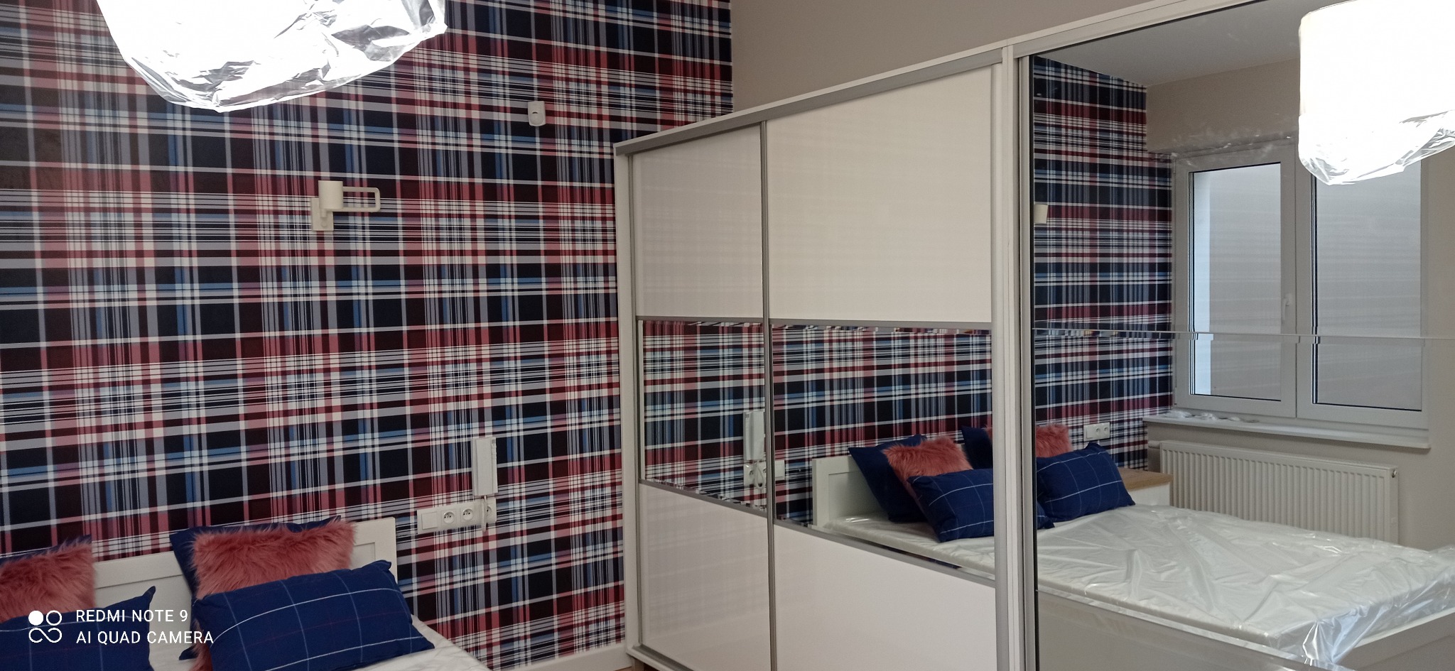 Pokój z tapetą w kratę, białym łóżkiem z niebieskimi poduszkami i szafą przesuwną z lustrami.