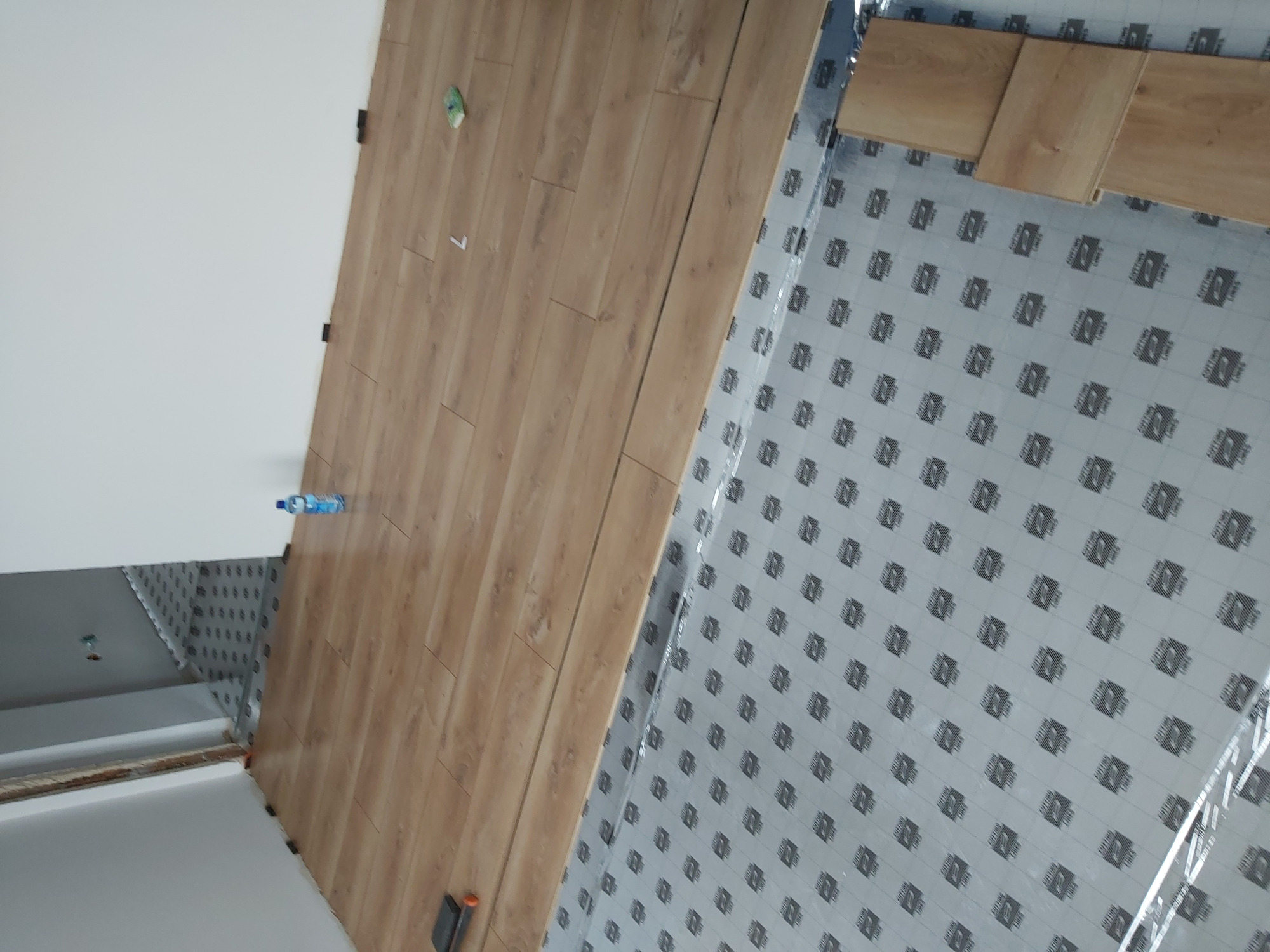 Fragment podłogi w trakcie montażu: ułożone panele w odcieniach brązu i przygotowane podłoże z folią izolacyjną z nadrukiem, widoczna butelka wody i narzędzia.